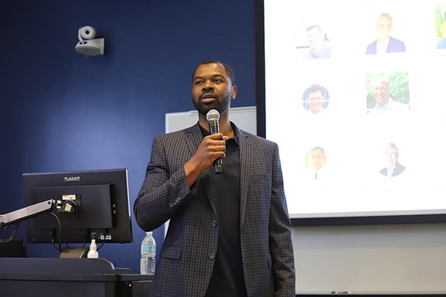 man wearing suit holding a mic while presenting a slideshow on screen behind him