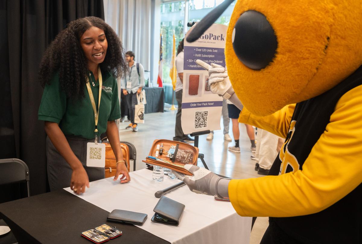 A Georgia Tech booth demonstration where a participant presents a wearable assistive device to Buzz and attendees at an innovation event.